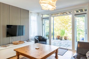 Ein modernes Wohnzimmer mit einem Flachbildfernseher, einem Holztisch, einer Ledersofa und einer Schiebeglastür, die zu einem Garten führt.