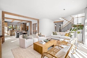 Ein modernes Wohnzimmer mit einer Sofagarnitur, einem Couchtisch und Rattan-Stühlen. Eine Treppe mit Geländer und eine Pflanze sind ebenfalls zu sehen.