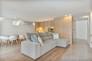 Un salon moderne avec un canapé d’angle, un coin repas doté d’une table en verre et une cuisine en arrière-plan.