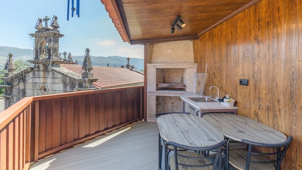 Un balcone con ringhiera in legno, un tavolo, due sedie, un angolo cottura con lavello e vista su un edificio storico con una torre dell'orologio.