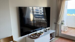 Een moderne woonkamer met een flatscreen televisie, een witte console tafel, een houten stoel en uitzicht op zee vanaf een balkon.