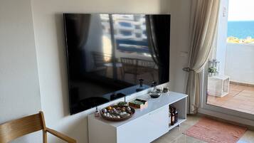 A modern living room with a flat-screen TV, a white console table, a wooden chair, and a view of the sea through a balcony.