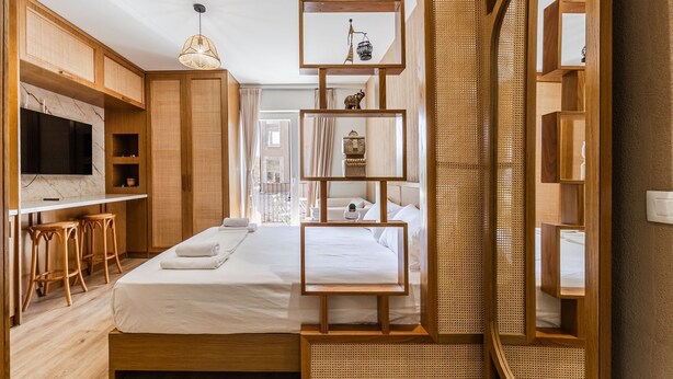 A modern hotel room with a wooden bed, a TV, a desk with stools, and a large mirror.