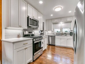 A modern kitchen with white cabinetry, stainless steel appliances, and a wooden floor. - Walk to OU - Spacious 4BR with Fire Pit (Norman)