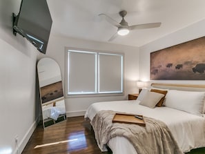 Ein Schlafzimmer mit einem Bett, einem Deckenventilator, einem an der Wand befestigten Fernseher, einem Fenster und einem Gemälde.