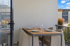 Une table à manger dressée avec des assiettes, des verres et des couverts, sur un balcon offrant une vue sur le ciel et les bâtiments environnants.