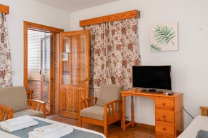 A room with a bed, a desk with a computer, two chairs, a television, and a door leading to an outdoor area.