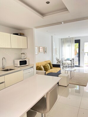 A modern kitchen with white cabinets, a microwave, and a sink. There is a dining area with a table and chairs, and a sofa with cushions.