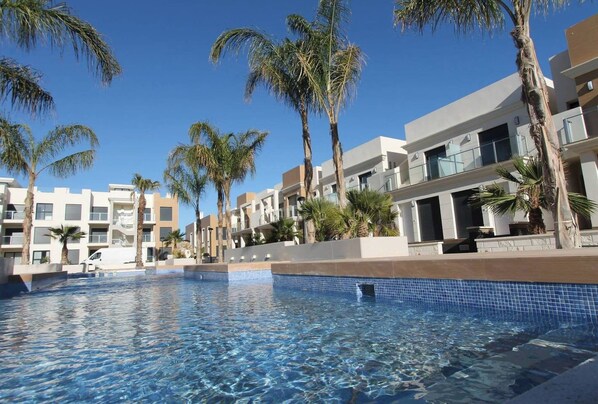 A swimming pool with clear blue water, surrounded by palm trees and modern buildings with balconies.