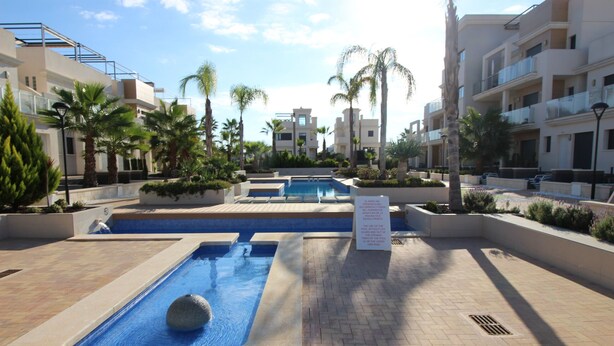 A swimming pool area with a rectangular pool and a smaller circular pool, surrounded by palm trees and modern buildings. - Duplez Zenia Beach (Orihuela)
