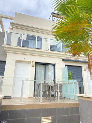 A modern two-story building with a balcony, a garage door, and a small outdoor seating area with chairs. - Duplez Zenia Beach (Orihuela)