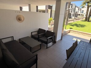 Un espace extérieur aménagé, abrité, avec des tables et des chaises, un chapeau accroché au mur, et une vue sur un jardin paysager et des bâtiments en arrière-plan.