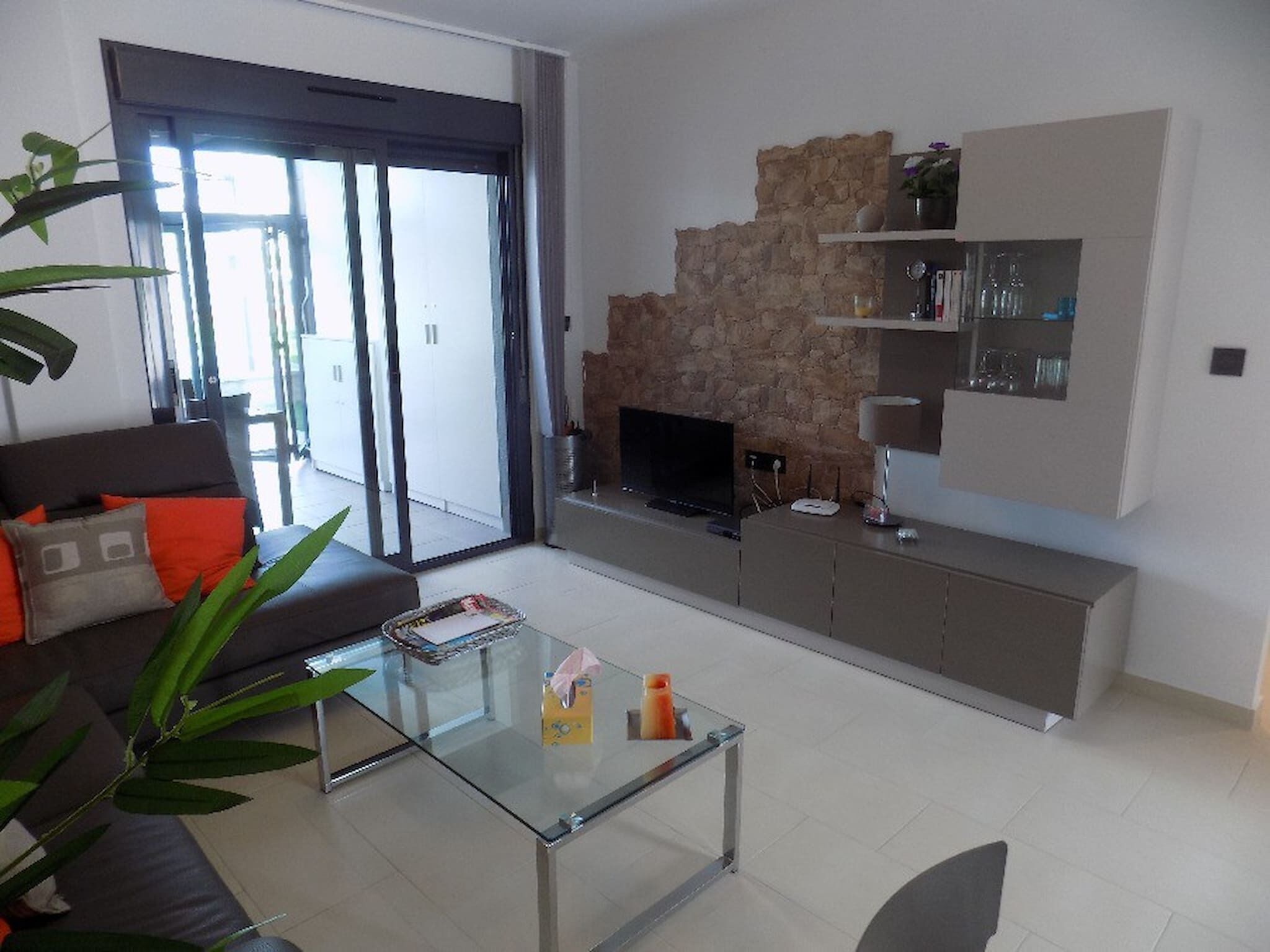 Uma sala de estar moderna com uma mesa de centro de vidro, um sofá com almofadas laranja, uma lareira de pedra e uma televisão.