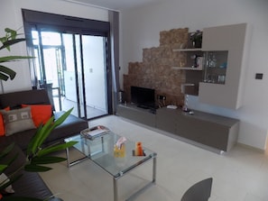 Una sala moderna con una mesa de centro de vidrio, un sofá con cojines anaranjados, una chimenea de piedra y un televisor.