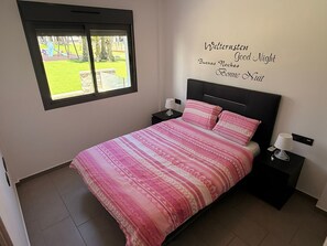 Ein Schlafzimmer mit einem Bett, zwei Nachttischen, einem Fenster und einer Wand mit der mehrsprachigen Aufschrift 'Good Night'.