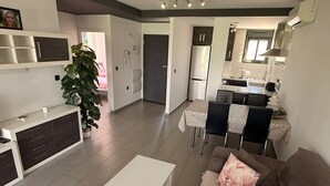 Uma sala de estar moderna com uma mesa de centro branca, um sofá marrom com almofada floral, uma área de jantar com mesa e cadeiras, e uma cozinha com geladeira e armários.
