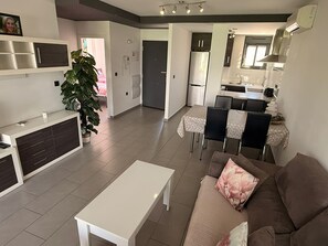 Ein modernes Wohnzimmer mit einem weißen Couchtisch, einem braunen Sofa mit einem floralen Kissen, einem Essbereich mit Tisch und Stühlen sowie einer Küche mit Kühlschrank und Schränken.