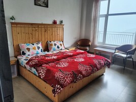 A bedroom with a wooden bed, a red floral bedspread, a chair, a small table, and a window with a view.