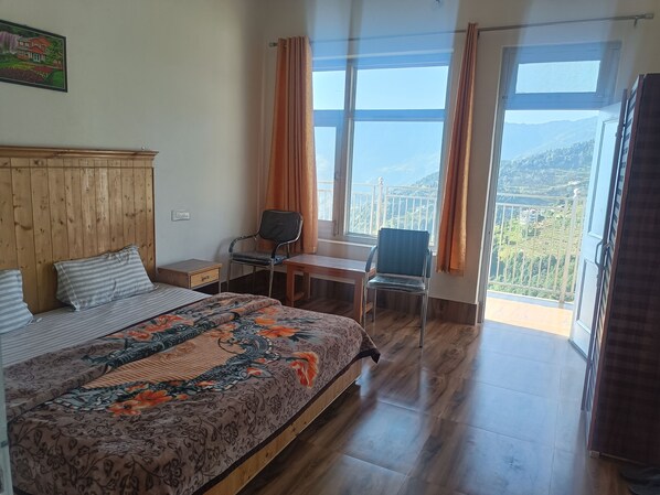 A bedroom with a bed, a desk, two chairs, and a view of a mountainous landscape through large windows.