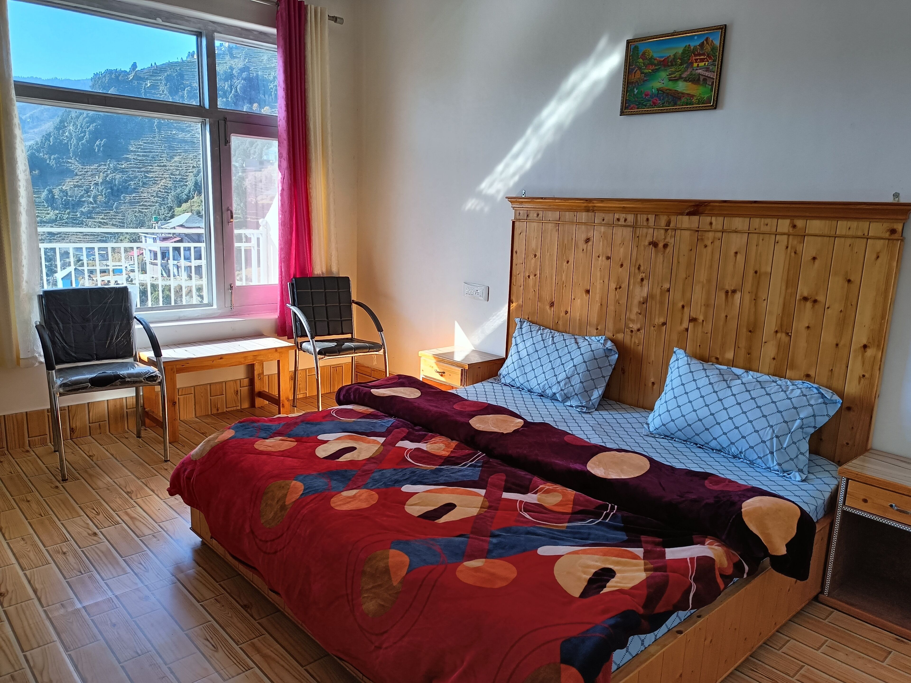A bedroom with a wooden headboard, a bed with patterned bedding, a desk with a chair, and a window with a view of a mountainous landscape.