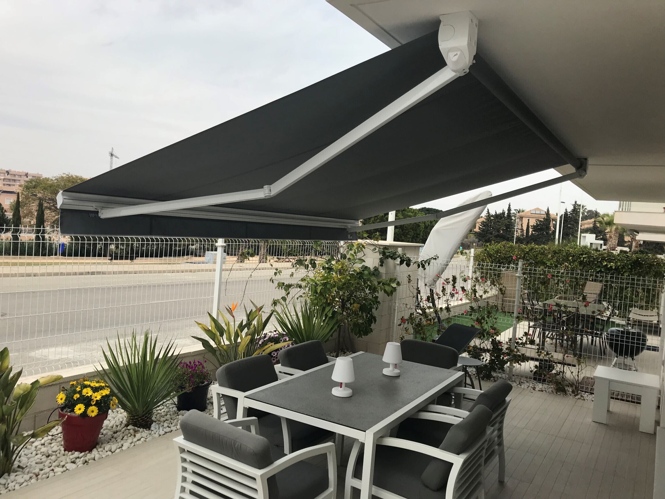 An outdoor patio with a table and chairs, surrounded by potted plants and a view of a street.