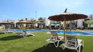 A poolside area with white lounge chairs and thatched umbrellas, a clear blue pool, and modern buildings in the background. - Groundfloor Tindra Punta Prima (Torrevieja)