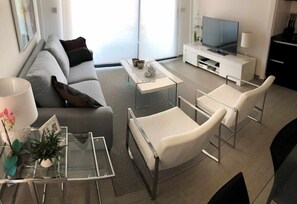 A modern living room with a sofa, two white chairs, a glass coffee table, a TV on a white stand, and a plant on a glass table. - House Tindra Homes Punta Prima (Torrevieja)