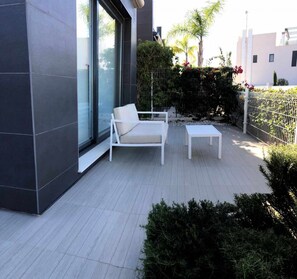 A modern outdoor patio with a white sofa and a small table, surrounded by greenery and a metal fence. - House Tindra Homes Punta Prima (Torrevieja)