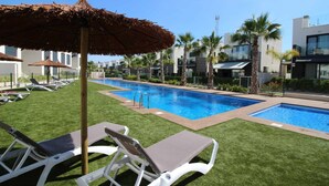 A swimming pool area with lounge chairs and umbrellas, surrounded by palm trees and modern buildings. - House Tindra Homes Punta Prima (Torrevieja)