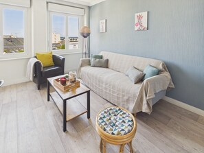 Ein Wohnzimmer mit einer Couch, einem Sessel, einem Couchtisch und einem kleinen Hocker. Ein Fenster bietet Blick auf Gebäude, und eine Stehlampe steht im Raum.