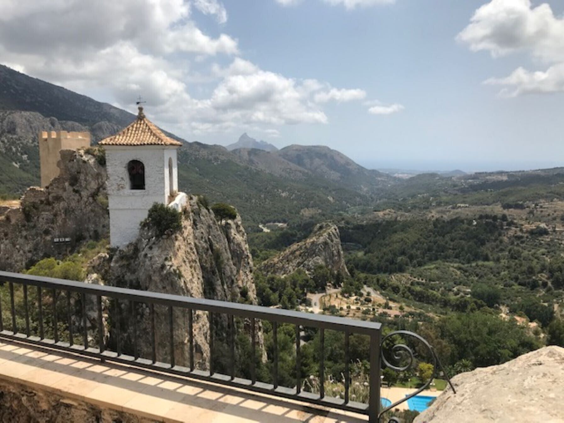 A white building with a red-tiled roof on a rocky outcrop, overlooking a valley with greenery and distant mountains.