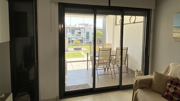A modern living room with a sofa, a dining table with chairs, and a sliding glass door leading to an outdoor patio area. - Groundfloor Oasis Beach Punta Prima IX (Torrevieja)