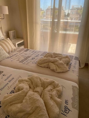 Ein ordentlich bezogenes Bett mit einem Bettlaken mit motivierenden Sprüchen, einer flauschigen weißen Tagesdecke und Blick auf Gebäude durch transparente Vorhänge.