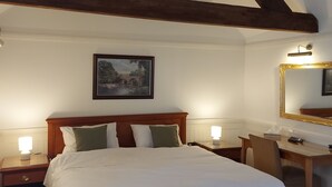 Un dormitorio con techo de madera, una cama con ropa blanca, dos mesitas de noche con lámparas, un escritorio con una silla y un cuadro en la pared.