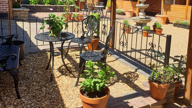 Una pérgola de madera con mesas y sillas, rodeada de plantas en macetas y un muro de ladrillo.