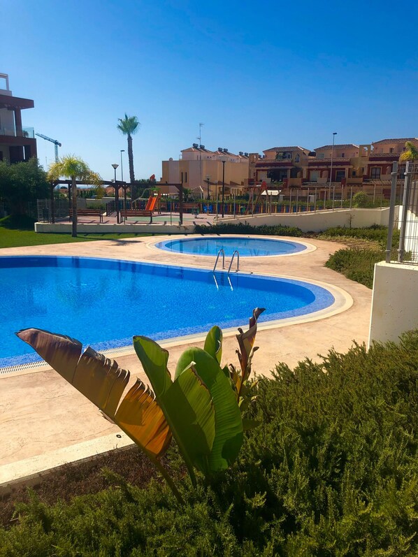 A swimming pool with two distinct sections, surrounded by greenery and a playground.