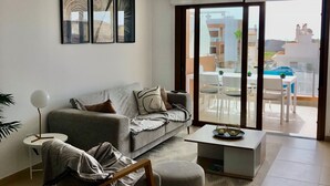 Ein modernes Wohnzimmer mit grauem Sofa, Couchtisch und Beistelltisch mit Lampe. Ein großes Fenster bietet Blick ins Freie.