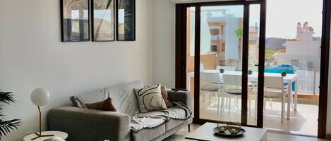 A modern living room with a grey sofa, a coffee table, and a side table with a lamp. There is a large window with a view of the outside.