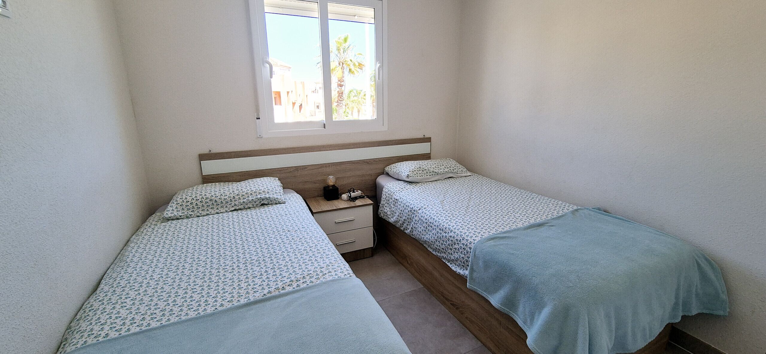 Une petite chambre avec deux lits simples, une table de chevet et une fenêtre donnant sur des palmiers.