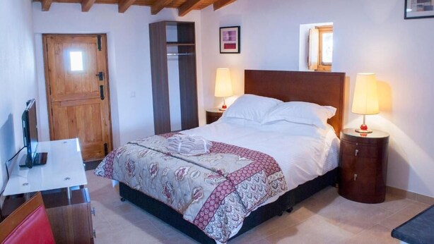 A bedroom with a wooden ceiling, a bed with a patterned bedspread, two bedside lamps, a wooden wardrobe, and a small table with a chair. - Casa De Campo De Oliveiras (Penela)