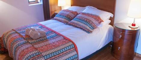 A neatly made bed with patterned bedding, a bedside table with a lamp, and a window with curtains.