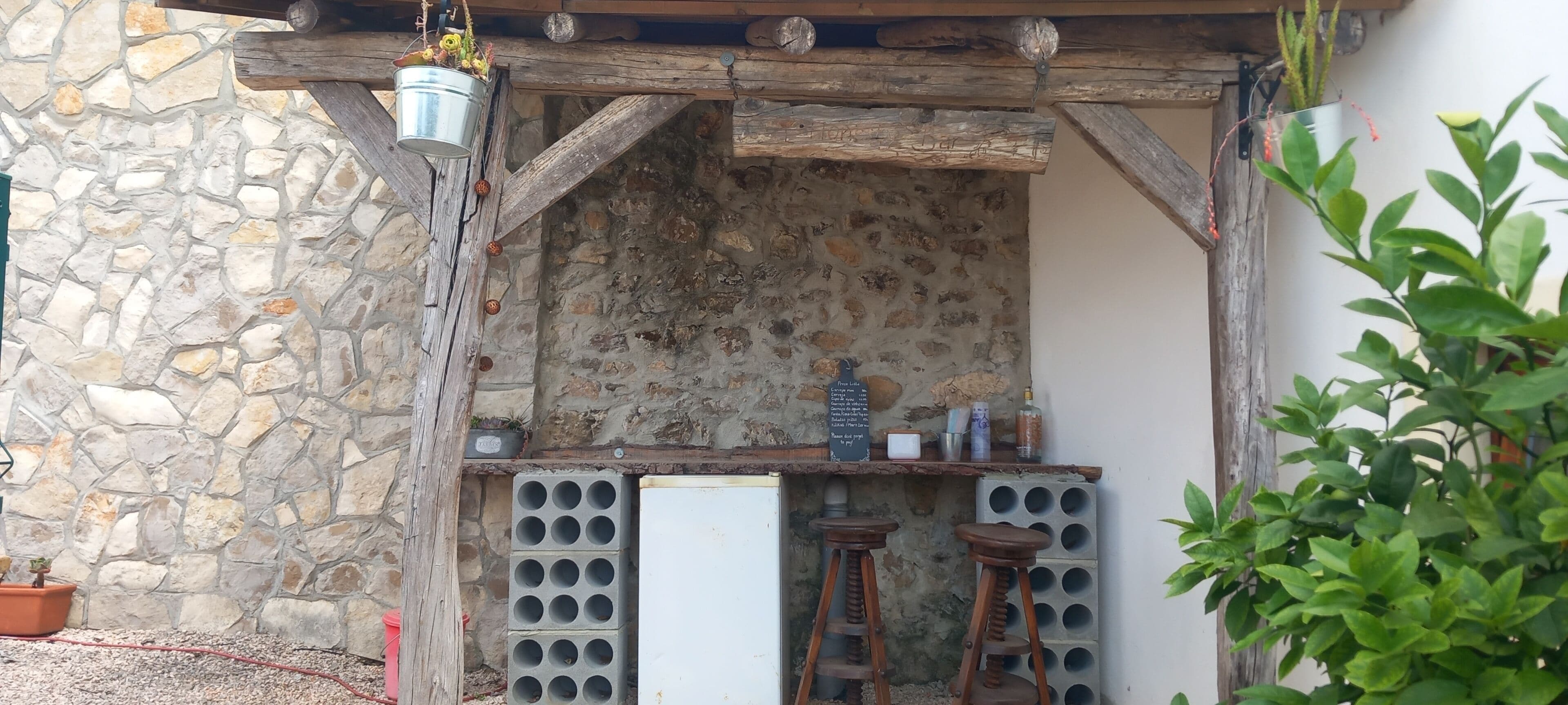 A rustic outdoor bar area with a stone wall, wooden beams, and a chalkboard menu.