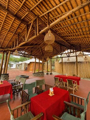 Un área de comedor al aire libre con mesas y sillas, techo de paja y vista de chozas tradicionales al fondo.