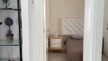 A bedroom with a bed, bedside table, and a nightstand. There is a shelf with decorative items on the left.