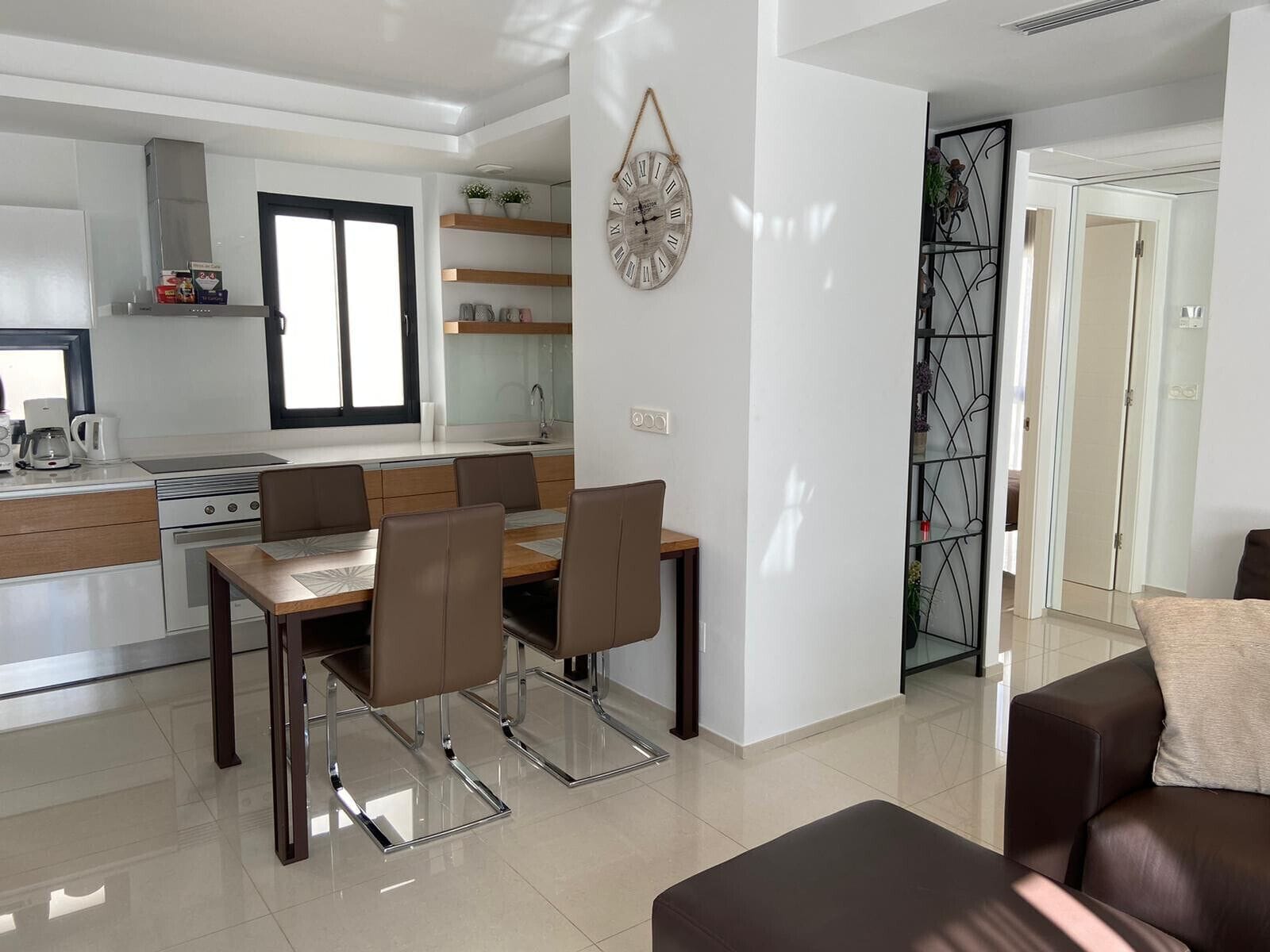 A modern kitchen with a dining area, a living room with a sofa, and a decorative wall clock.