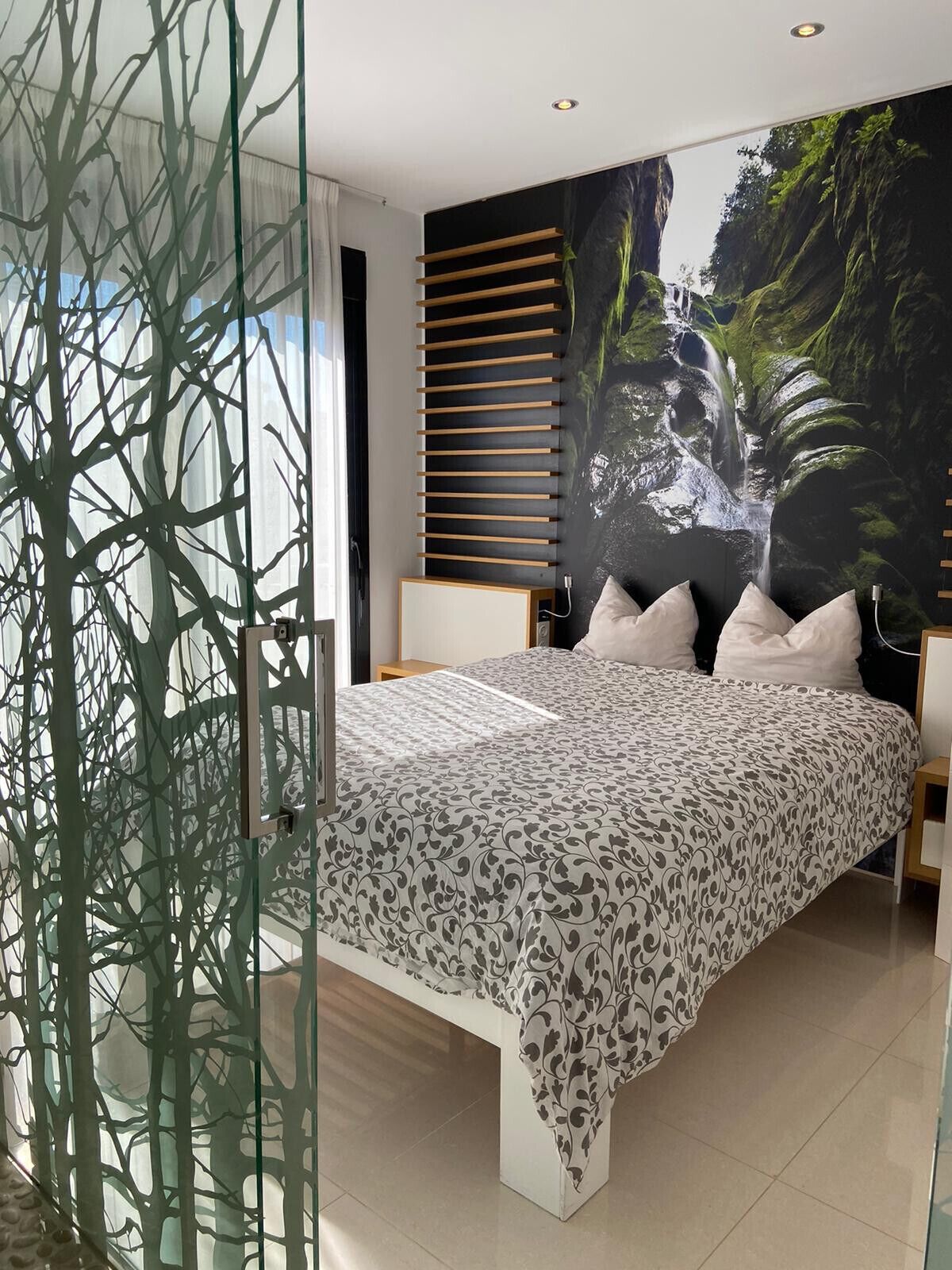 A modern bedroom with a large bed, a glass partition, a wall-mounted shelf, and a large nature-themed mural.