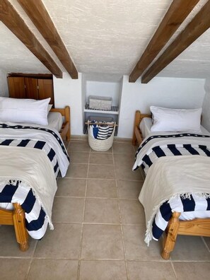 Una habitación con dos camas, cada una con colcha a rayas, cabeceros de madera y almohadas blancas. Hay una pequeña cesta de almacenamiento en el centro de la habitación.