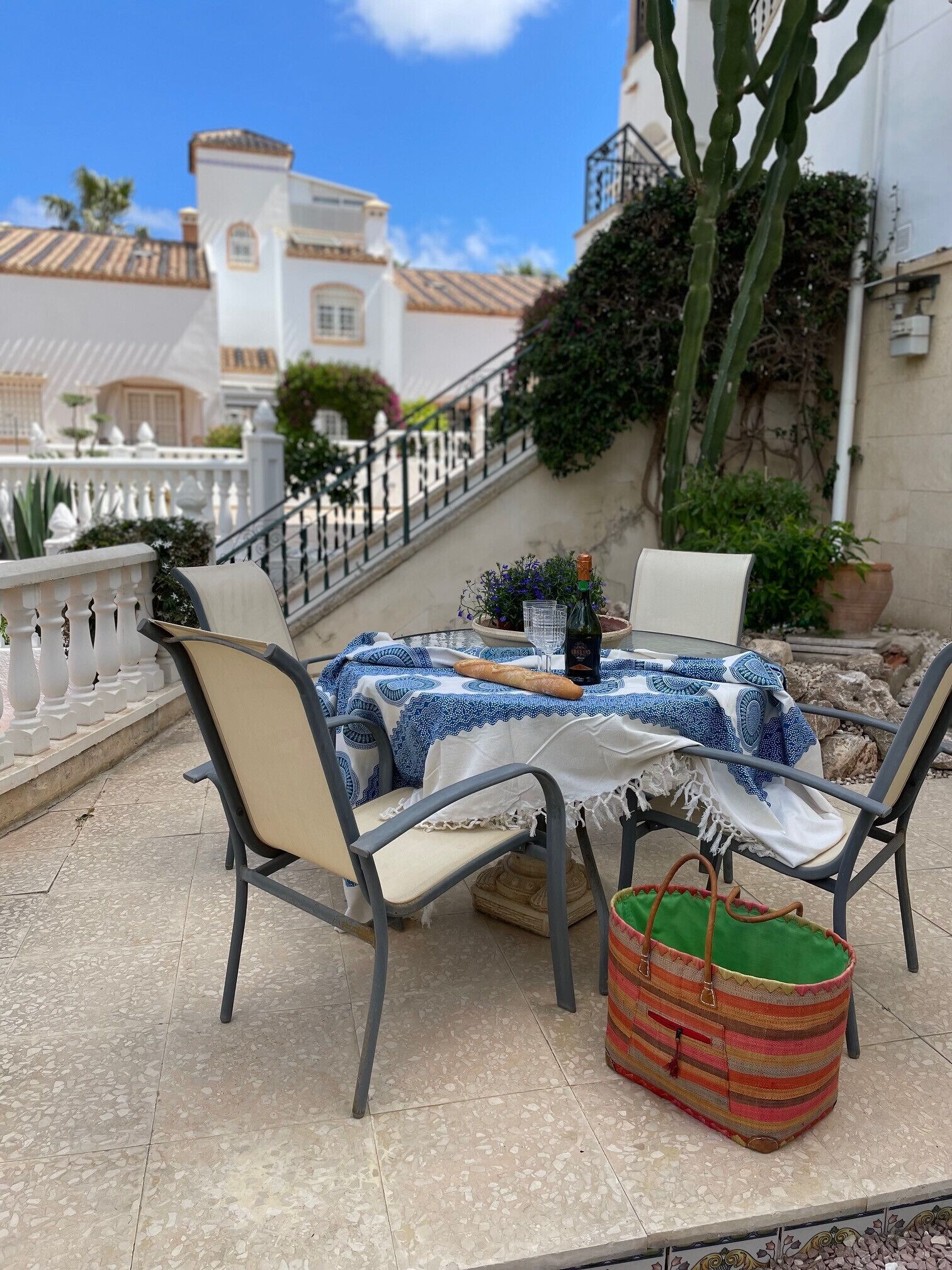 An outdoor patio with a table set for two, chairs, a basket, and a view of a building with a staircase.