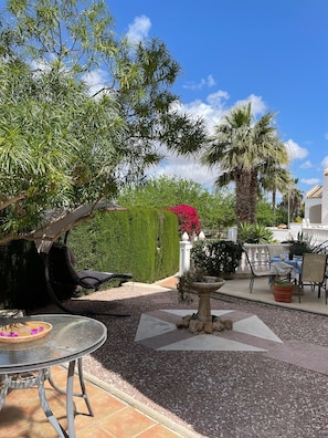 Eine Terrasse mit einem runden Tisch, Stühlen, einer Hängematte, einem kleinen Brunnen und verschiedenen Pflanzen und Bäumen.