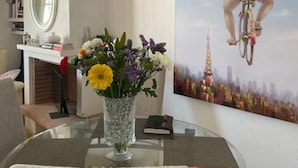 A dining area with a glass table, chairs, a vase of flowers, a painting of a person on a bicycle, and a bookshelf. - Penthouse Villamartin Laplaza Destino (Orihuela)